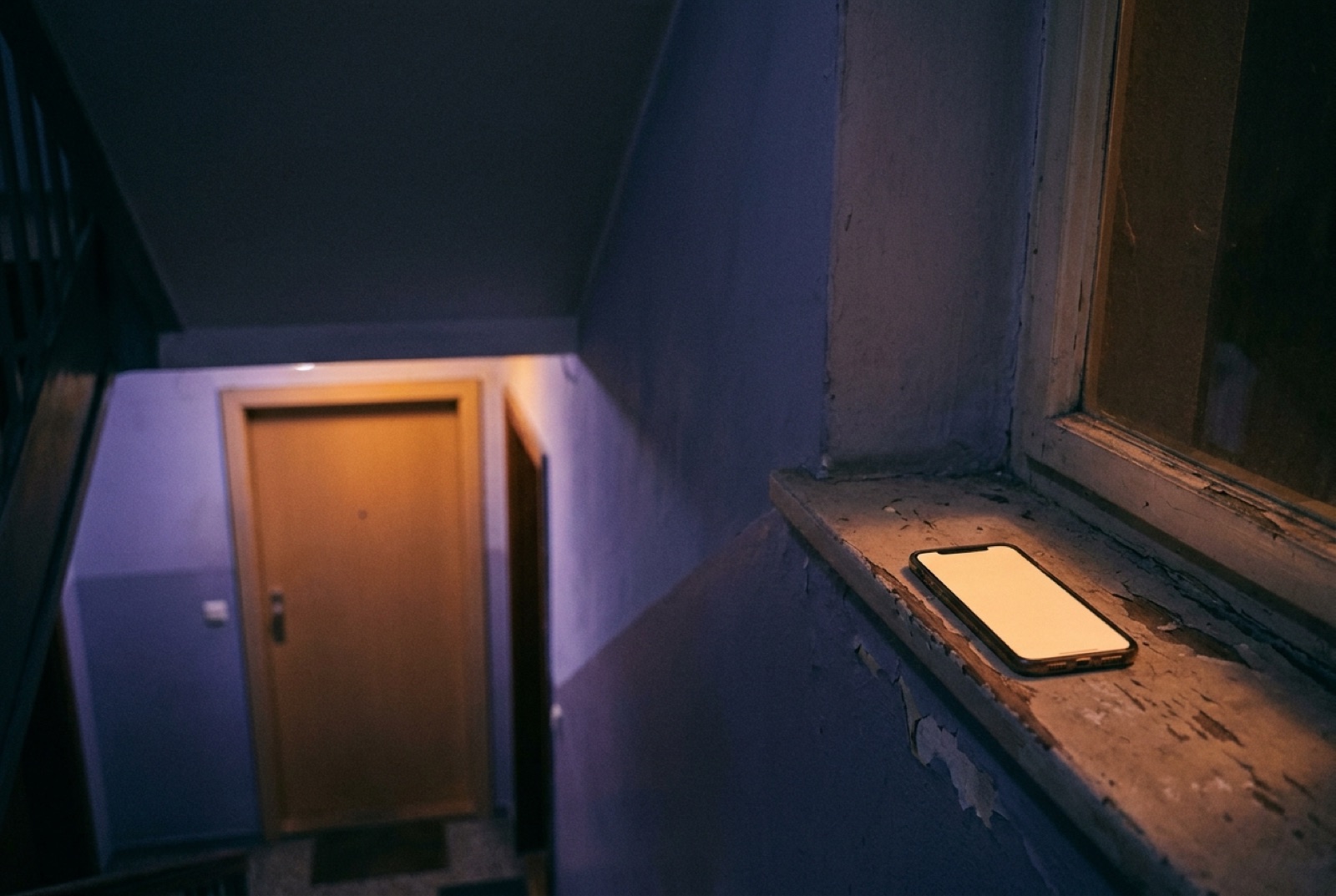 Ein Telefon leuchtet auf der Fensterbank im Treppenhaus, dahinter eine warm erleuchtete Wohnungstür.