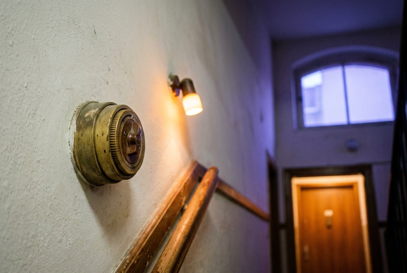 Ein alter Treppenhaus-Zeitschalter aus Messing an der Wand, daneben eine warme Wandleuchte.