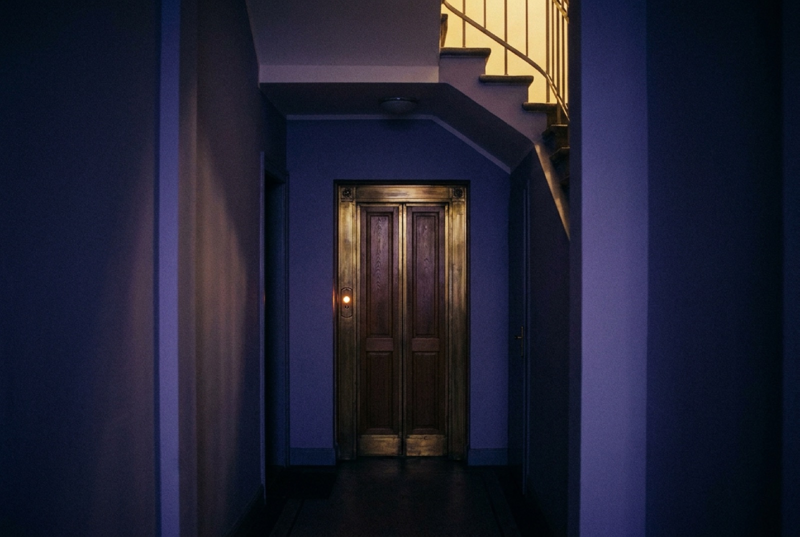 Geschlossene Aufzugtür im Treppenhaus eines Altbaus bei Nacht, der Rufknopf leuchtet warm
