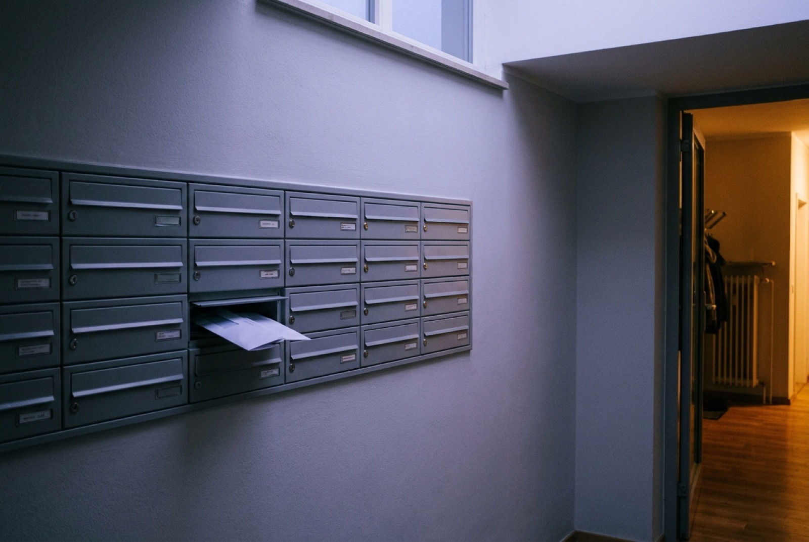 Briefkastenanlage im Hausflur, ein Fach leicht geöffnet, warmes Licht aus einer Tür