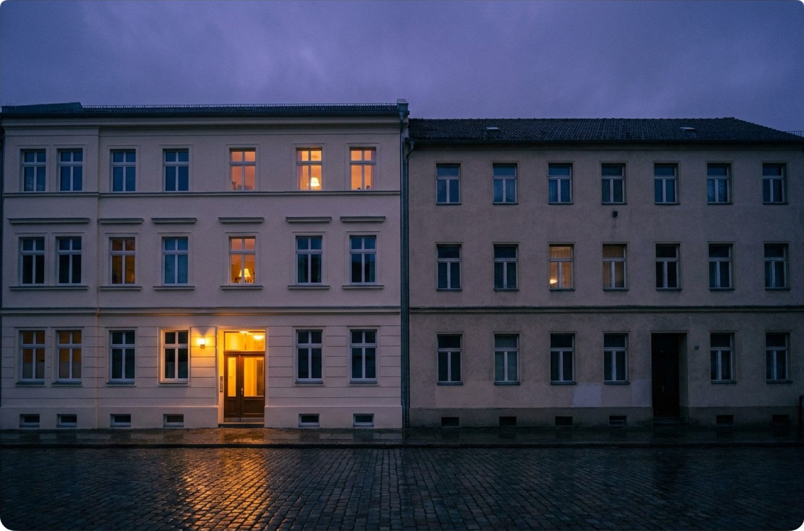 Zwei Mehrfamilienhäuser nebeneinander in der Dämmerung, eines frisch renoviert und warm erleuchtet